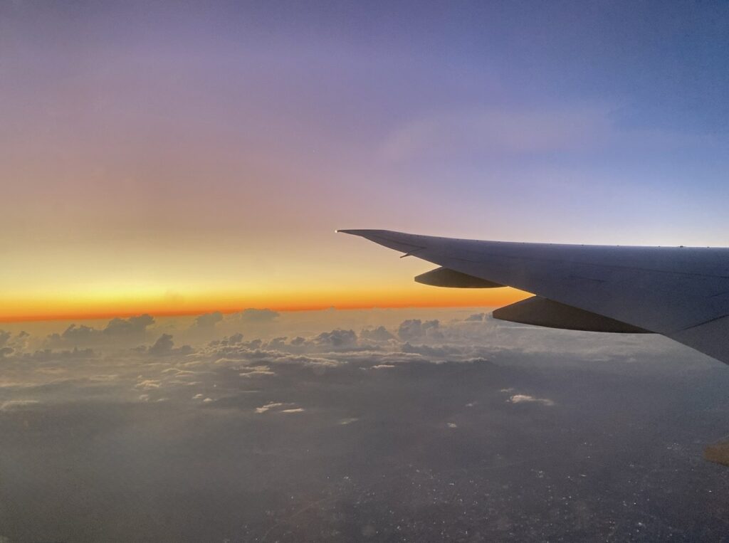 飛行機の窓から見た朝焼けの空と雲海。やわらかなグラデーションの空に、機体の翼が浮かぶ幻想的な風景